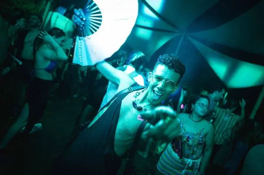 festivals in costa rica-11 | Pacific Photography Man in sunglasses and an open shirt dances joyfully at a crowded, neon-lit party. He wears a chunky necklace and suspenders, with others dancing in the background.