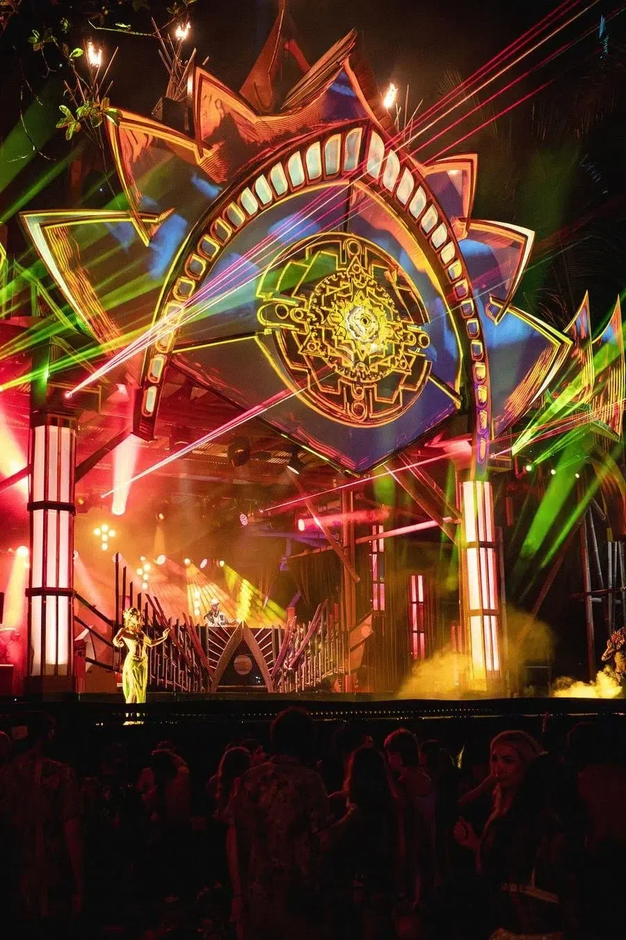 Colorful concert stage with bright laser lights, a neon geometric arch backdrop, and a performer on the left amid a crowd at night.