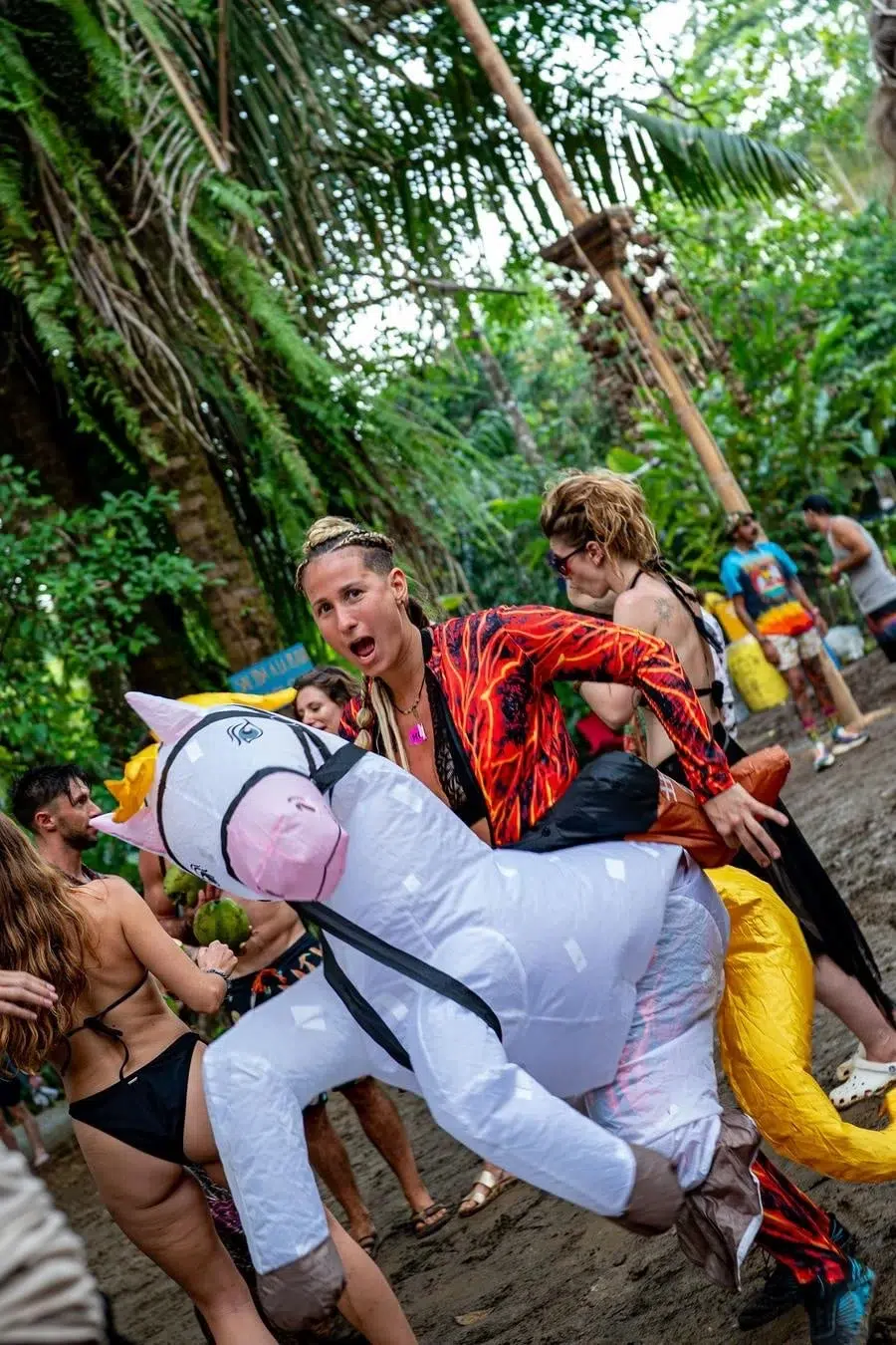 Person in a fiery red shirt rides a large inflatable white unicorn at an outdoor tropical party.