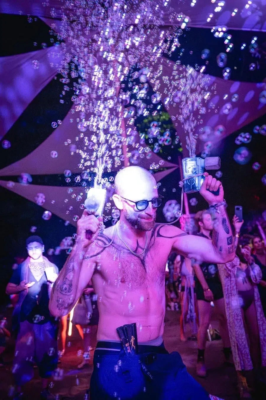 Shirtless, bald man with glasses sprays champagne at a purple-lit party, bubbles filling the air as a cheering crowd watches.