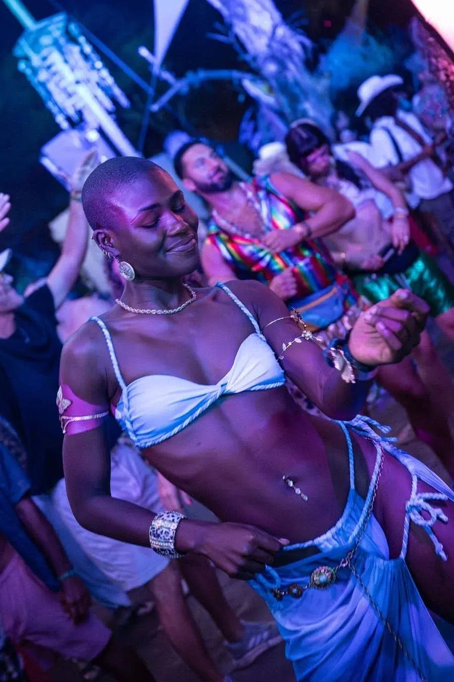 Person dancing at a crowded nightclub wearing a light blue bikini-style top and matching skirt with jewelry under colorful lights.