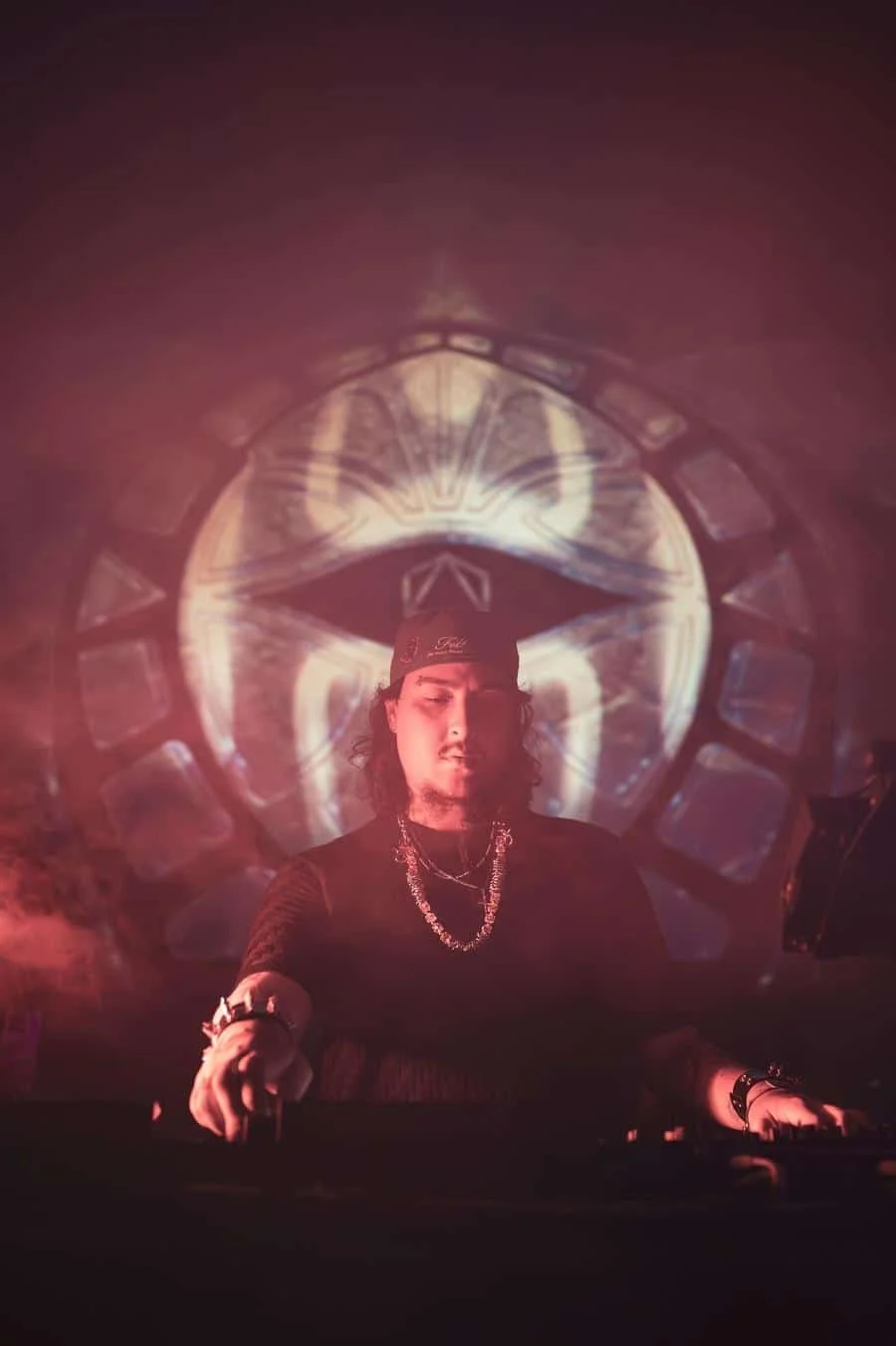 DJ wearing a cap and necklace mixes music on a dark stage with a circular, glow-filled backdrop behind him.