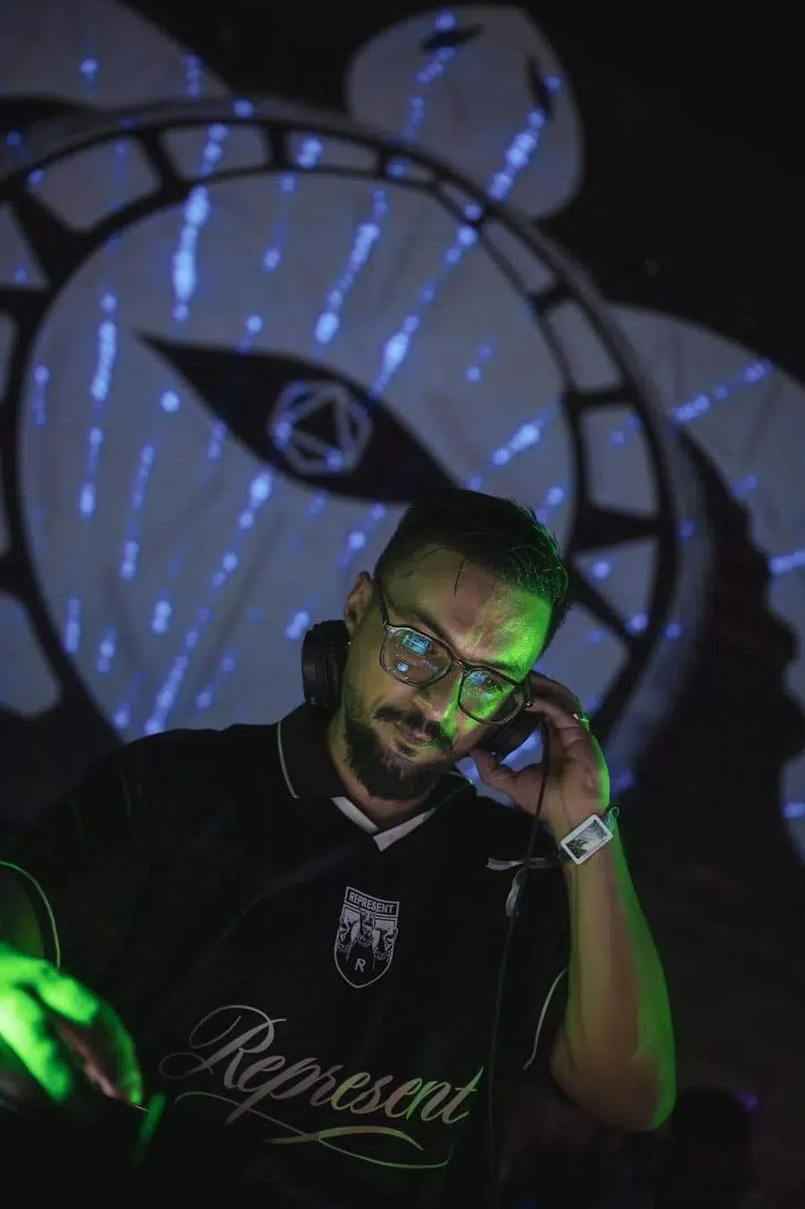 DJ wearing glasses and headphones outdoors at night, green light on face, large abstract projection in background.