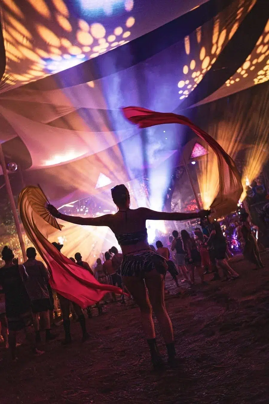 Dancer on a festival ground twirls red fabric under colorful stage lights, crowd watching in the background.