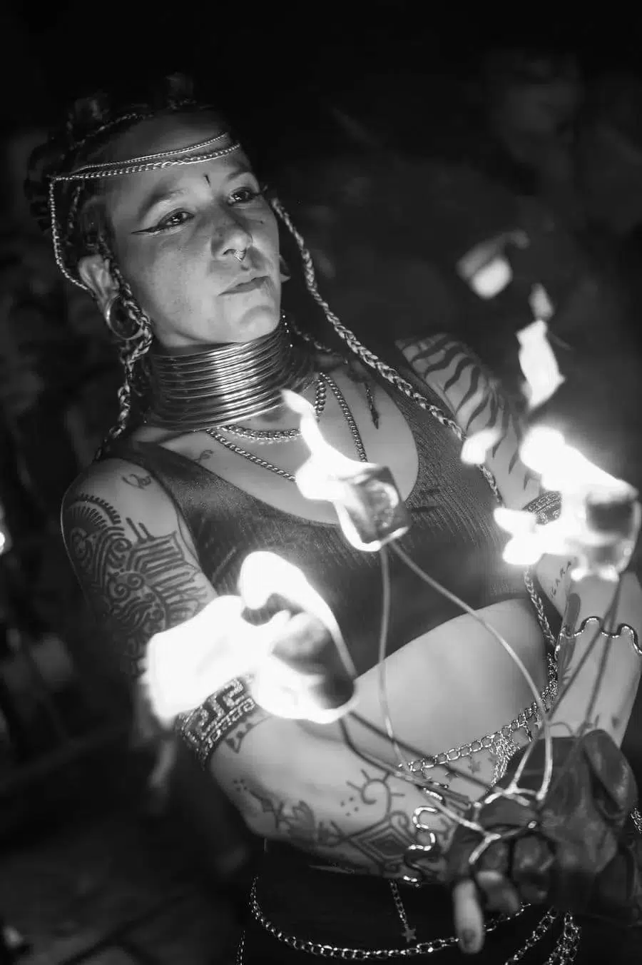 Black-and-white image of a tattooed fire performer with braids and thick neck rings, spinning flaming wicks.