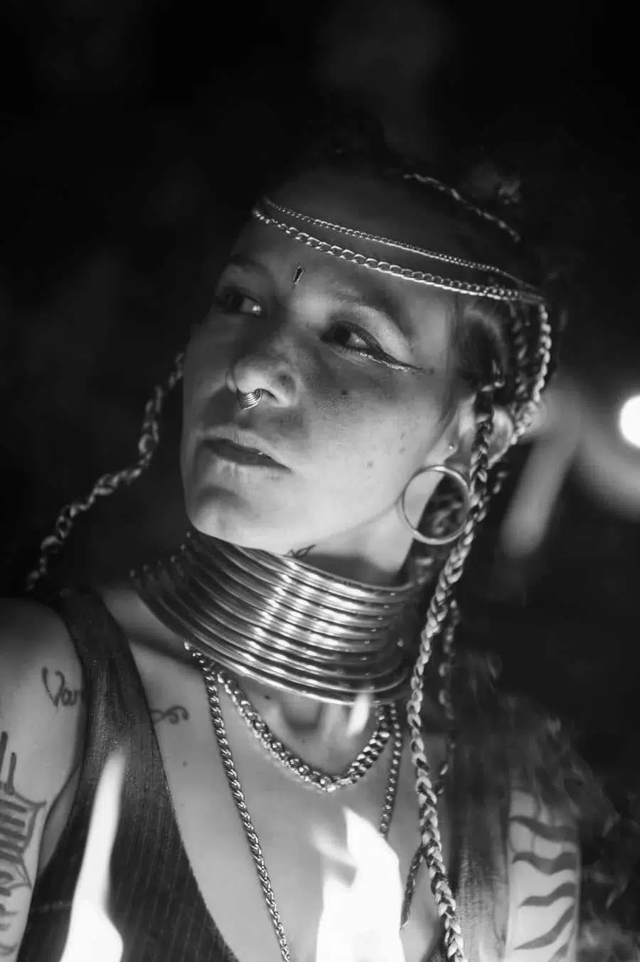 Portrait of a person with braids and stacked metal rings around the neck, nose ring, earrings, and tattoos in black-and-white lighting.