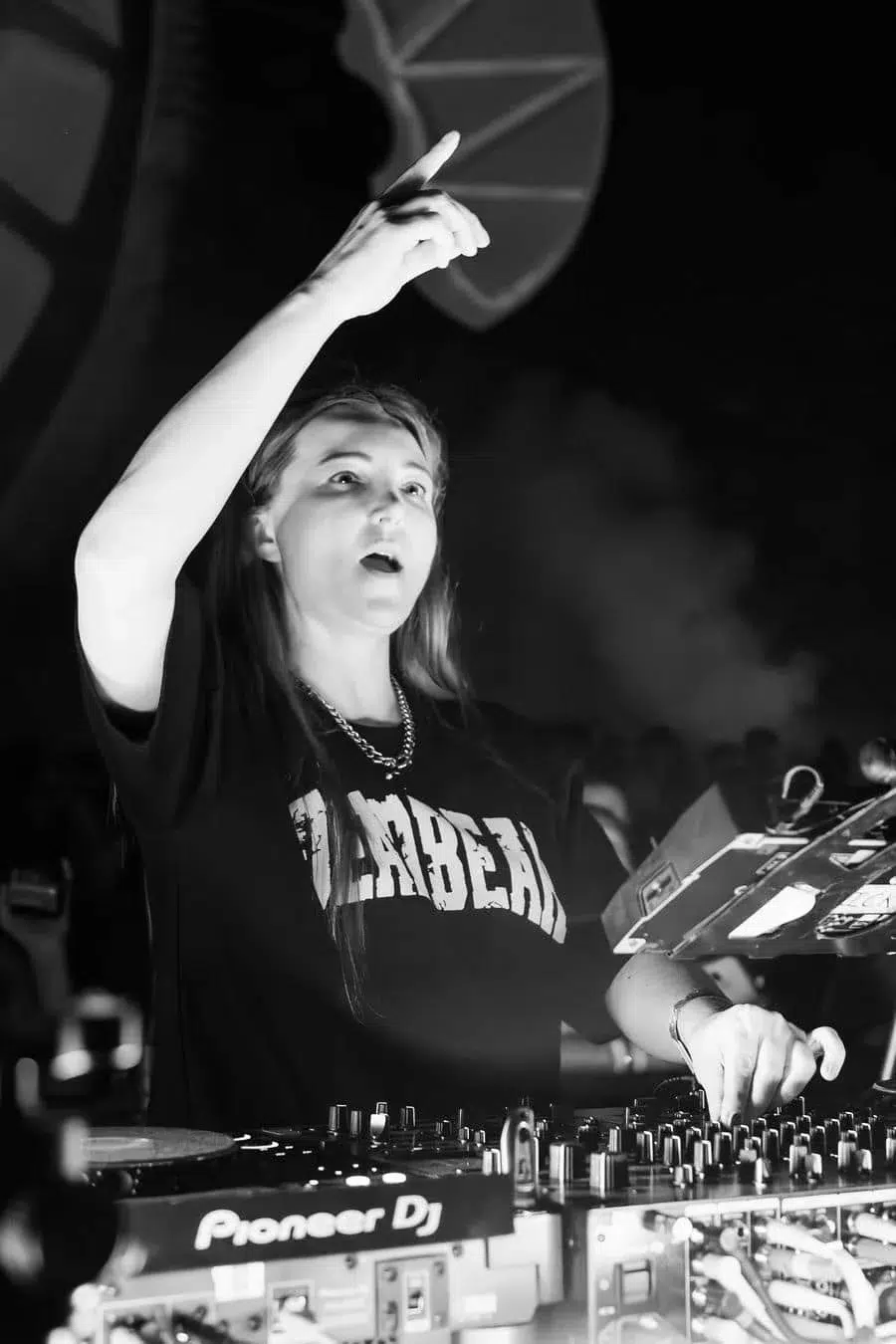 Black-and-white photo of a female DJ performing at a club, raising her right hand while adjusting the mixer labeled Pioneer DJ in front of her.