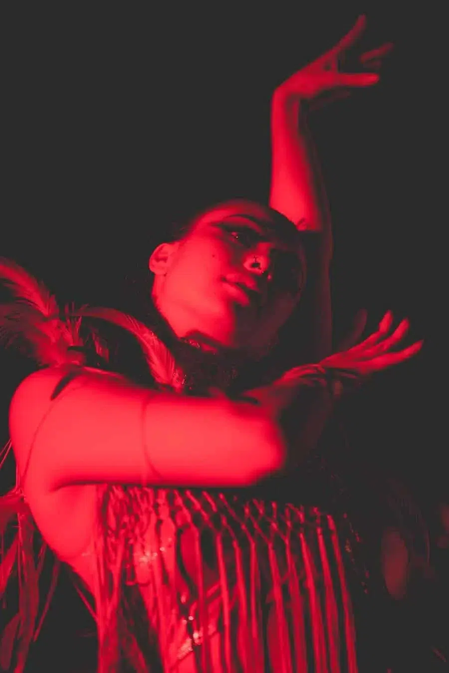 Dancer leans back under red stage lighting, fringe dress swaying as arms arc above.