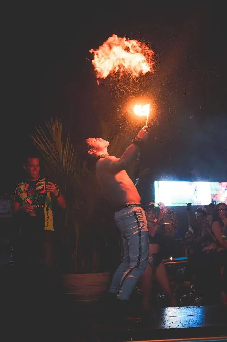 Male fire performer on a dark stage, raised torch producing a large flame while crowd watches.