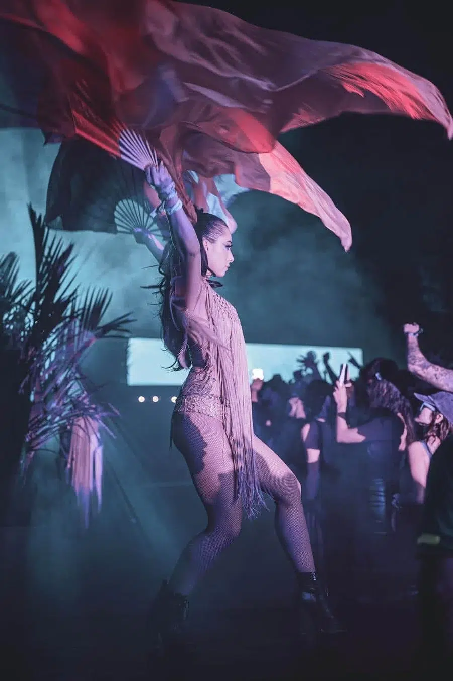 Dancer on a nightclub stage, arms raised, with a flowing red scarf and fringe outfit under purple lights, audience nearby.