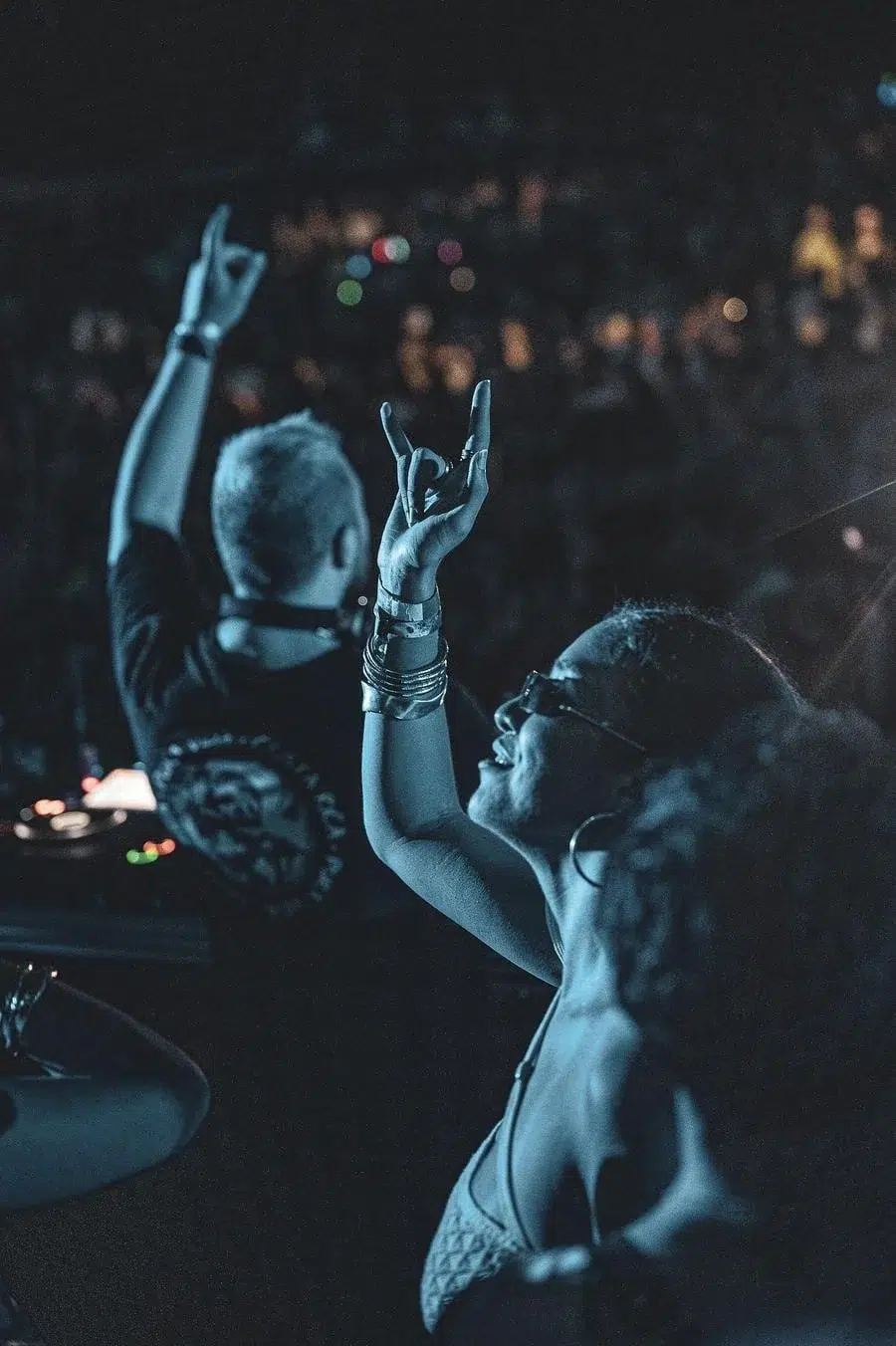 DJ on a dimly lit stage raises hands in the air as a blue-tinted club crowd cheers behind her.