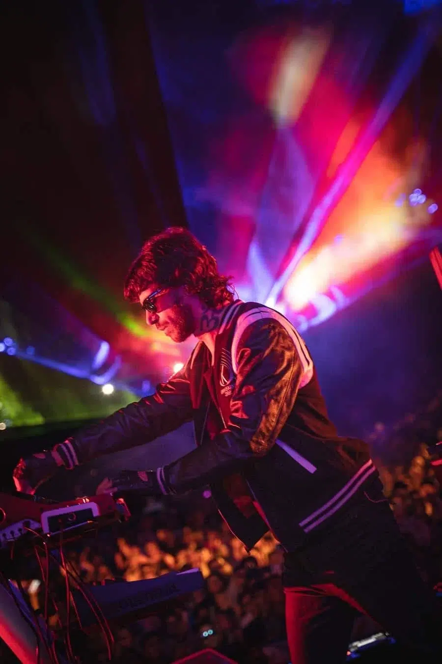 DJ wearing sunglasses and a varsity-style jacket, mixing on electronic gear on a colorful lit stage with a cheering crowd behind him