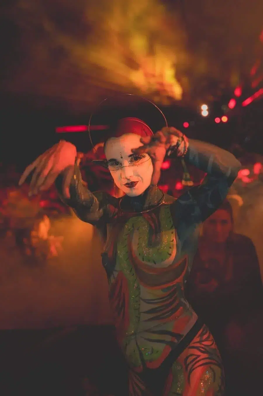 Performer with white face makeup in a colorful bodysuit, framing their face with a red hoop in a smoky, orange-lit venue.