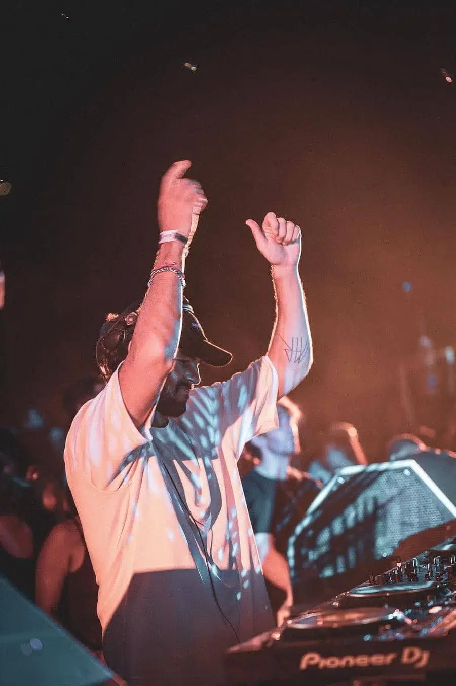 DJ with headphones and raised arms performing at a club, Pioneer DJ mixer in foreground, crowd in background.