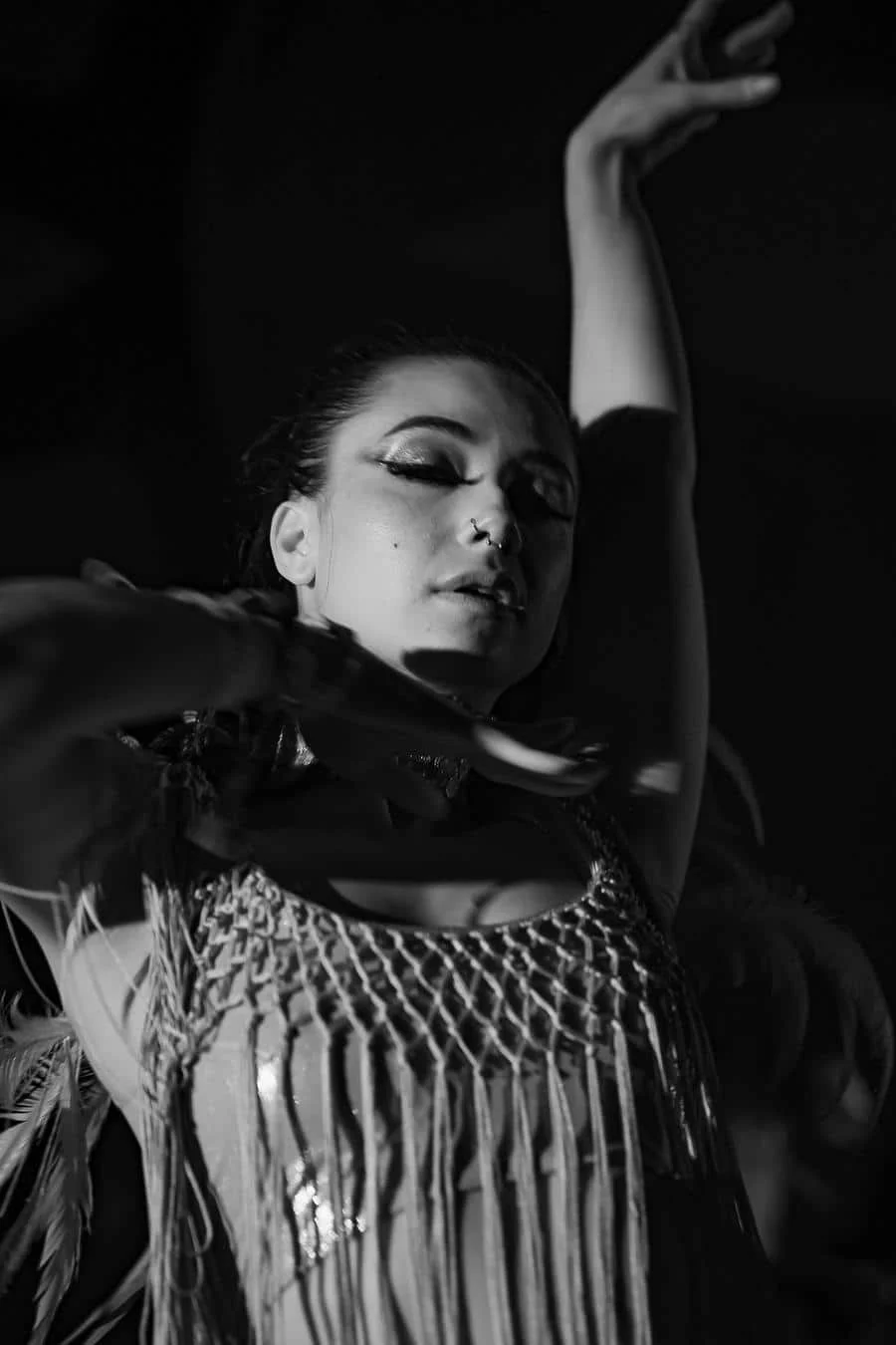 Female dancer in a fringe top raises one arm while closing her eyes, performing on stage in black and white.