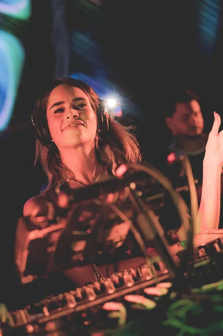 Female DJ wearing headphones and smiling while mixing on a DJ mixer in a dim club, colorful lights in the background.