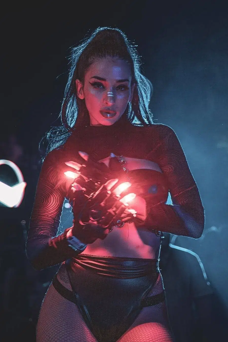 Female performer on stage wearing a black mesh outfit, holding glowing red LED lights with both hands amidst blue stage lighting