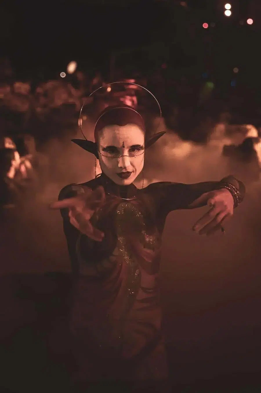 Performer in a dramatic fantasy costume with white face makeup and elf ears, reaching toward the camera under red smoky lighting and a halo ring behind the head.