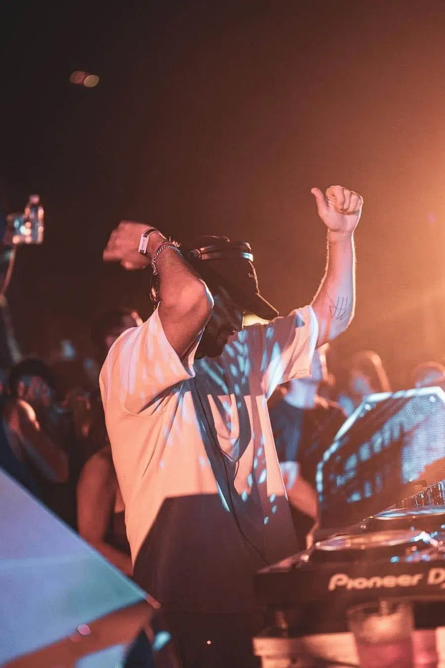DJ on stage at a dim club, wearing a cap and white shirt, arms raised as he spins records.