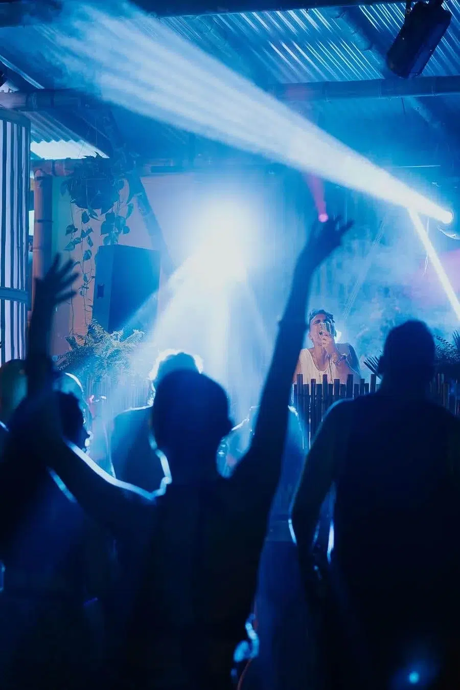 Crowd dancing with raised hands in a blue-lit nightclub as a DJ performs on stage with fog and spotlights behind them
