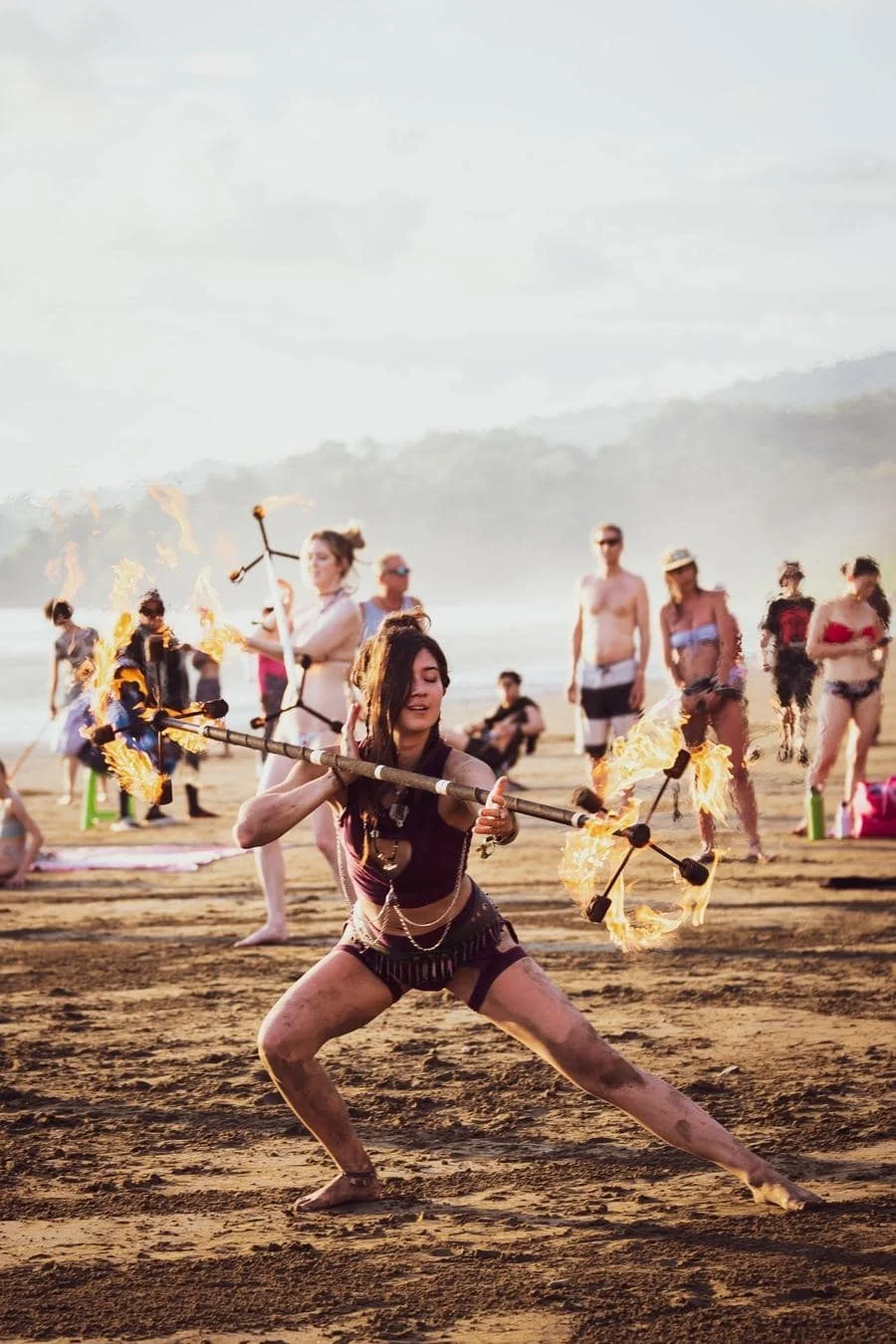 Woman performing a fire poi routine on a sandy beach with a cheering crowd watching in the background.