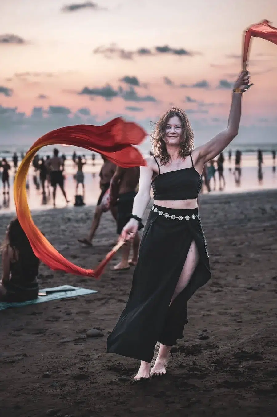 Woman dancing on a beach at sunset, twirling a red scarf with arms raised, wearing a black crop top and long skirt.