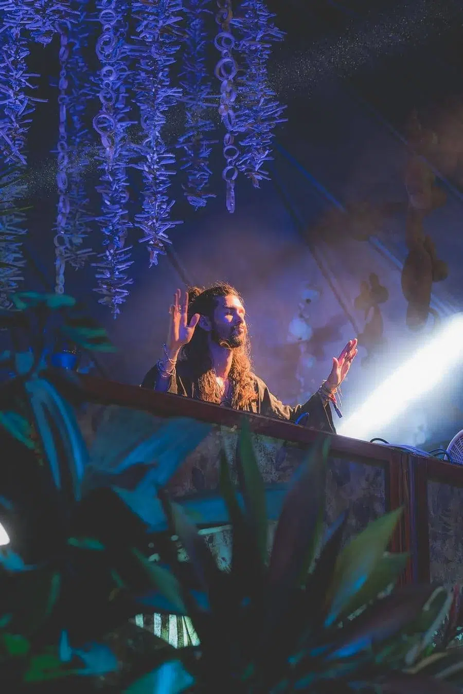 Bearded DJ with long hair on a balcony stage, hands raised, under blue-purple lights with hanging plants overhead.