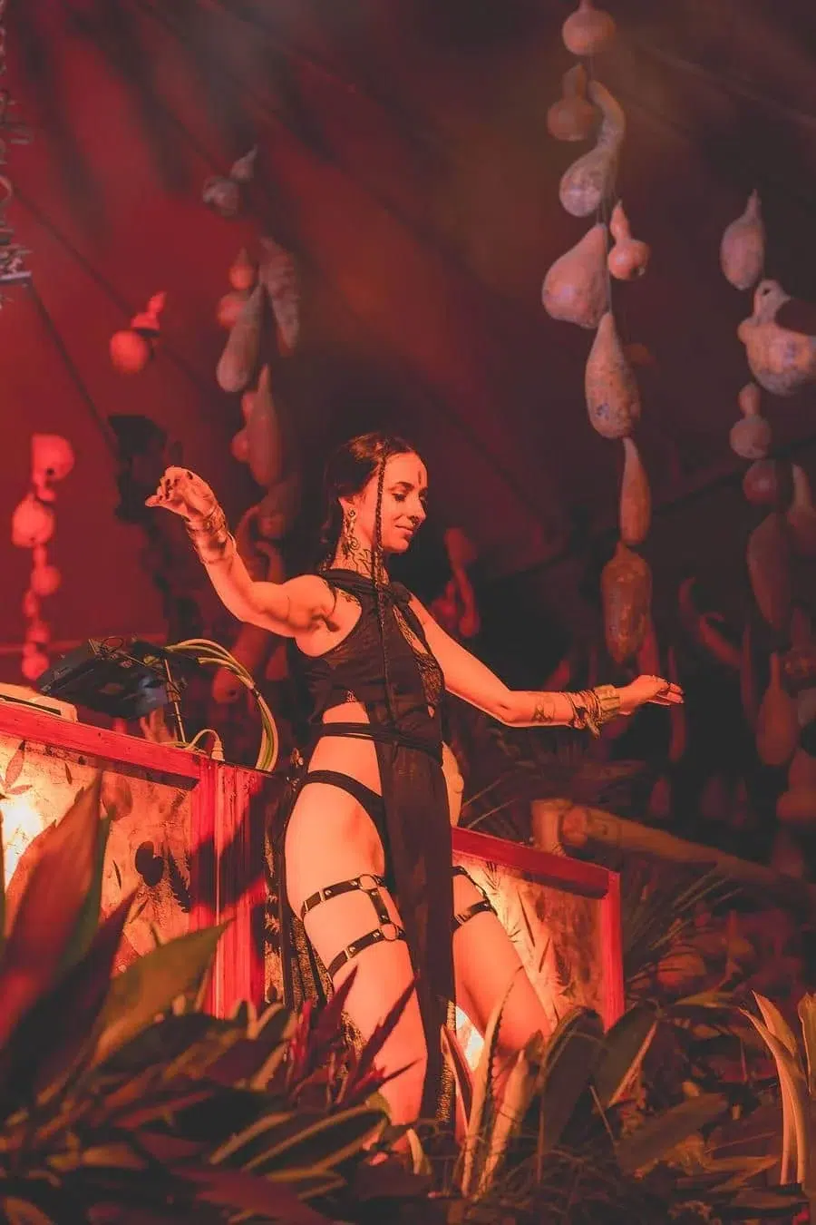 Dancer in a black cut-out costume performs on a red-lit stage, arms raised, with hanging gourds and tropical plants around.