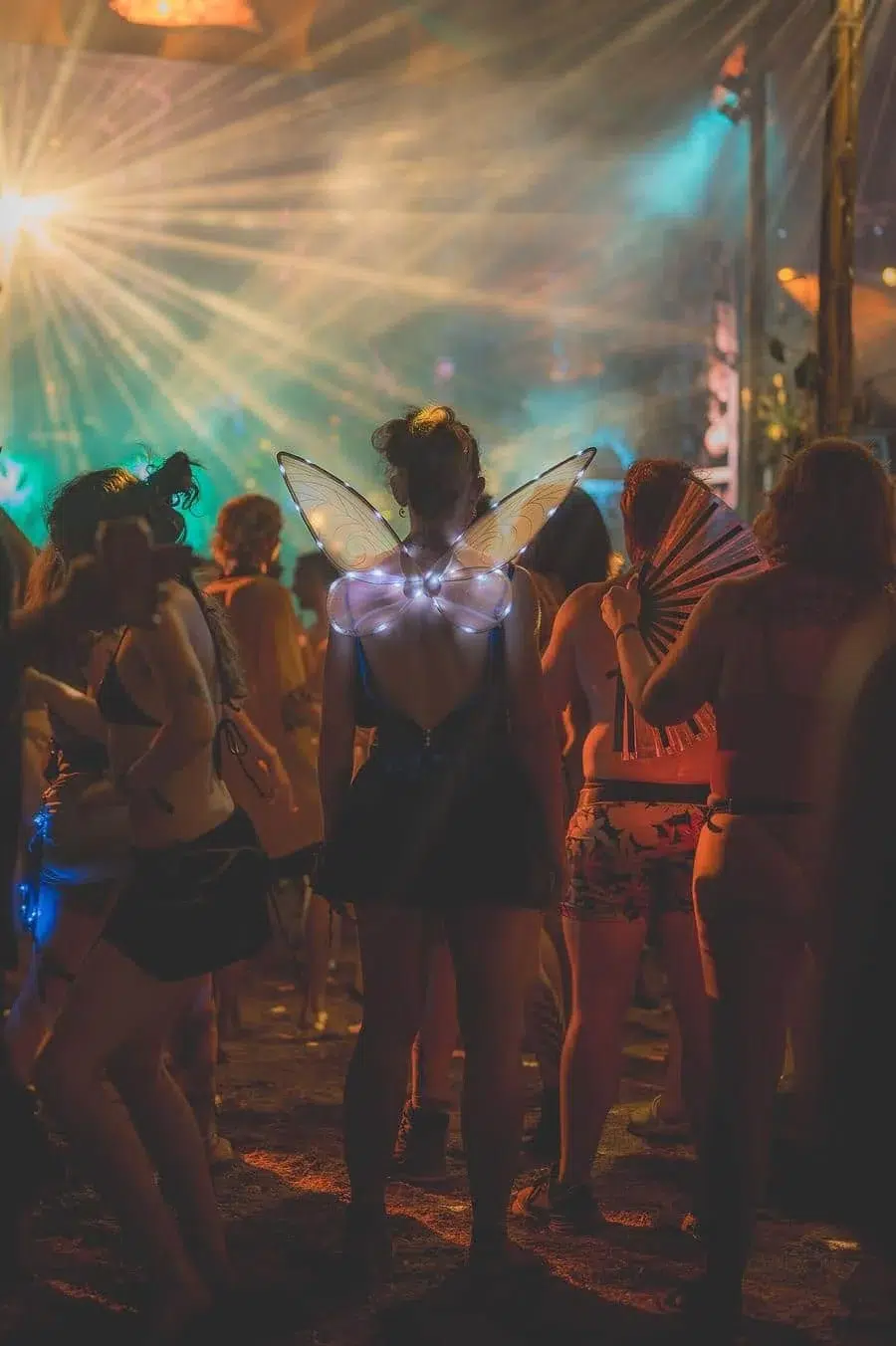 Person wearing glowing butterfly wings at a nighttime festival, among a crowd with colorful lights and fans.