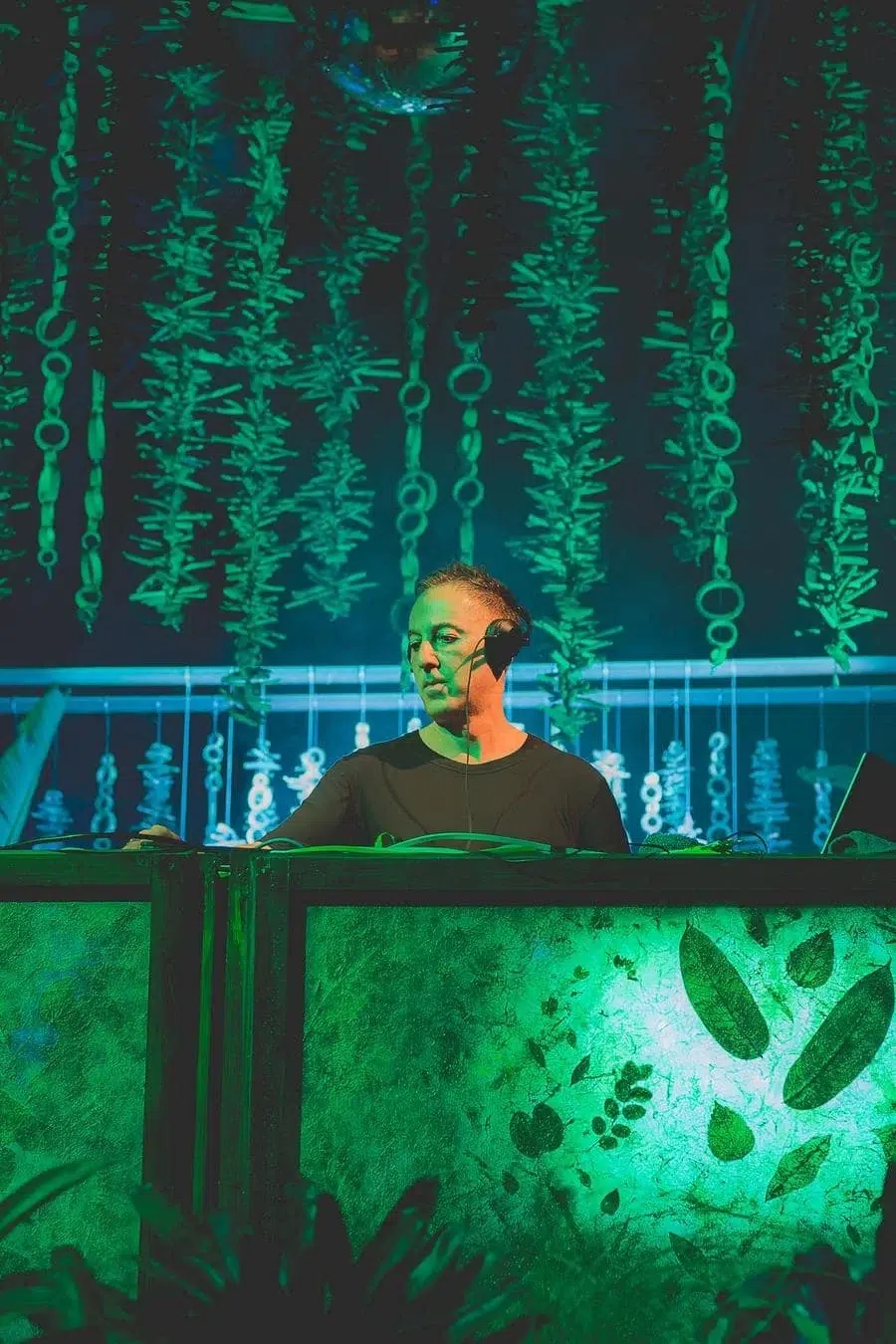 DJ performing at a green-lit club, wearing headphones behind a neon-green booth with hanging vines overhead.