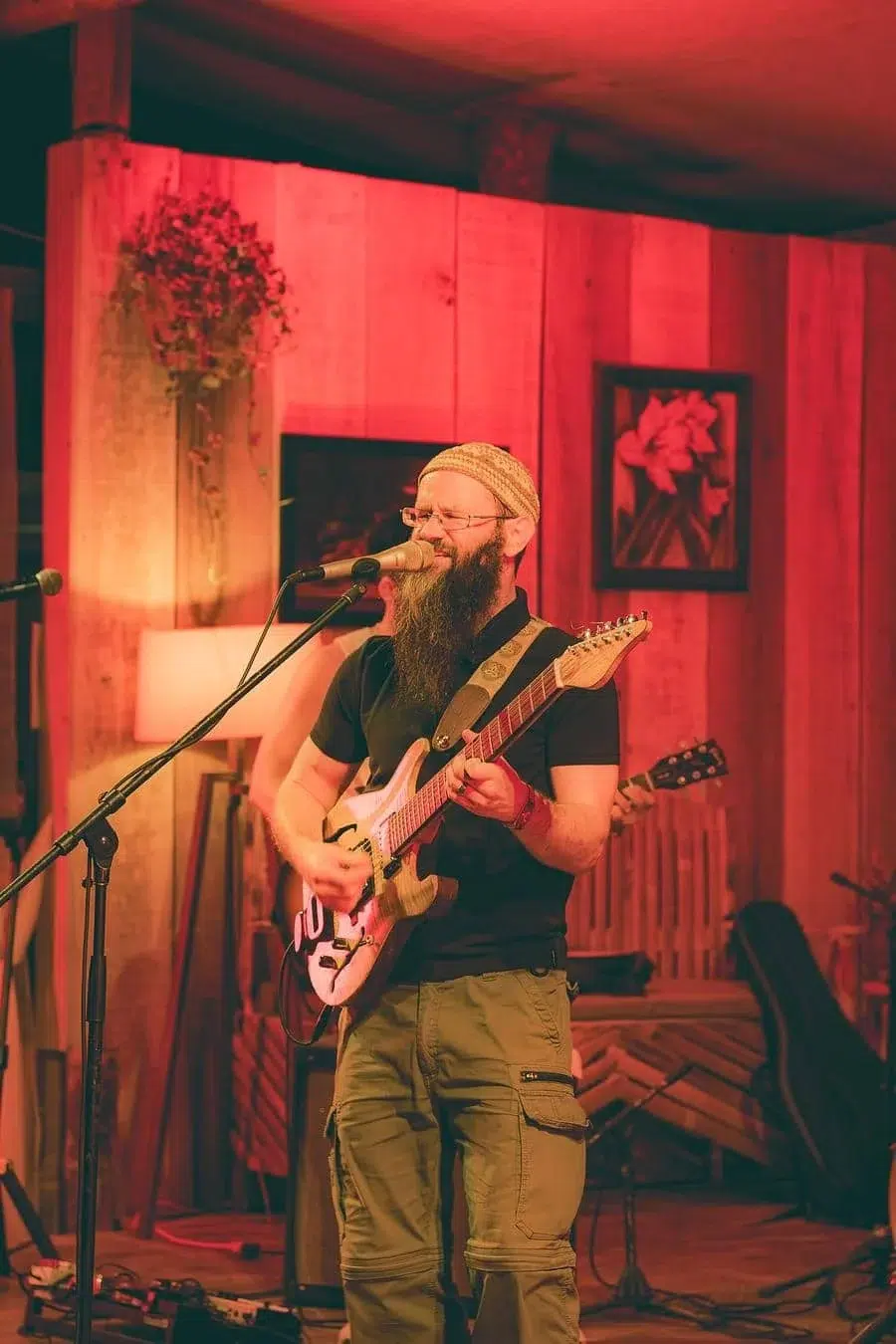 Bearded musician wearing a knit cap sings into a microphone while playing an electric guitar on a warmly lit indoor stage.