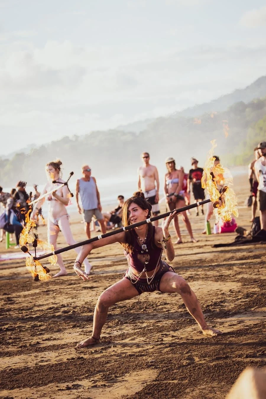 Female fire performer crouches on a sandy beach, spinning a flaming staff while a crowd watches in the background.