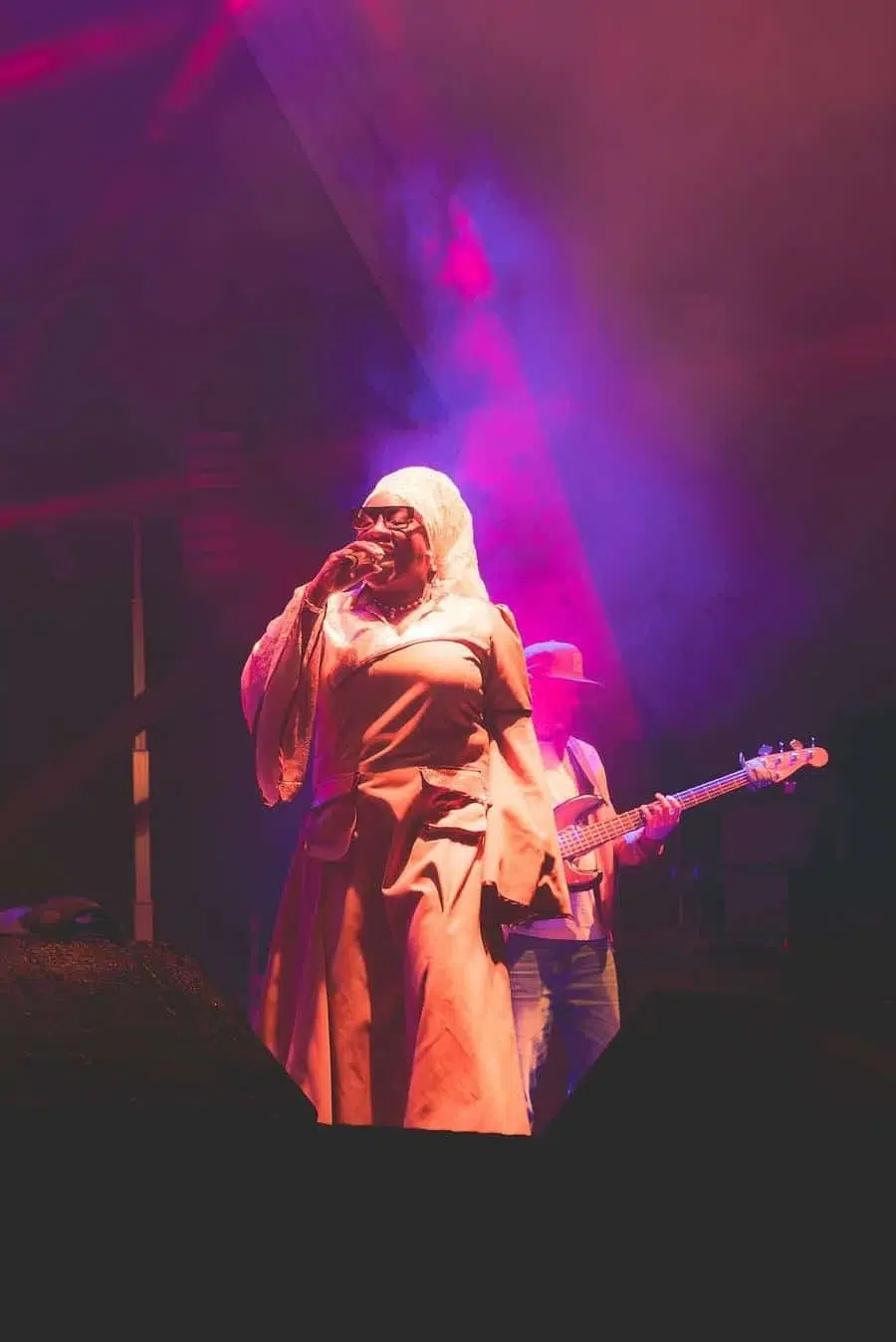 Female singer on stage in a long beige robe, wearing a head covering, singing into a microphone with a bassist in the background amid pink-purple stage lights.