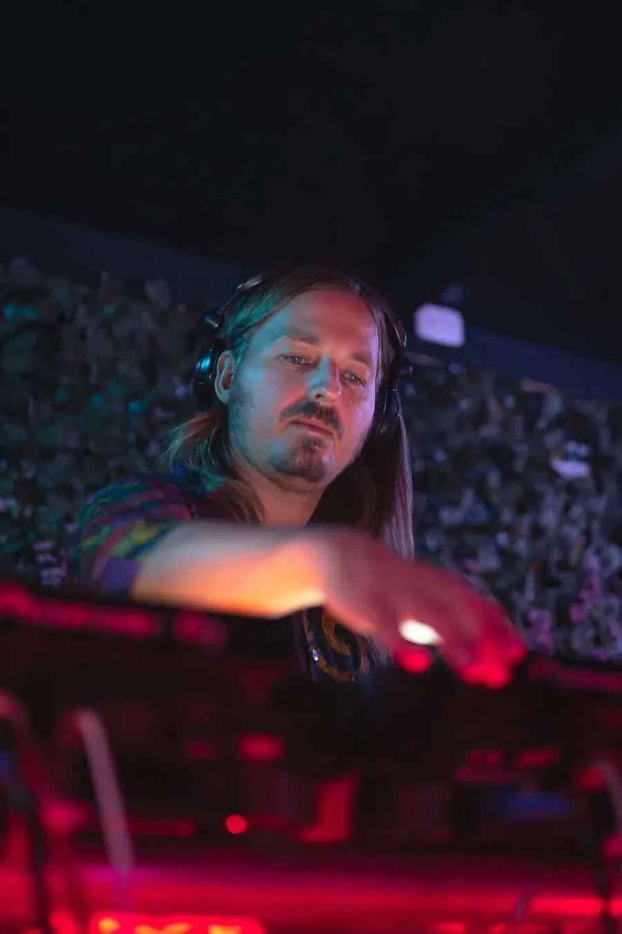 DJ with long hair wearing headphones mixes tracks at a club, colorful stage lights and a cheering crowd in the background.
