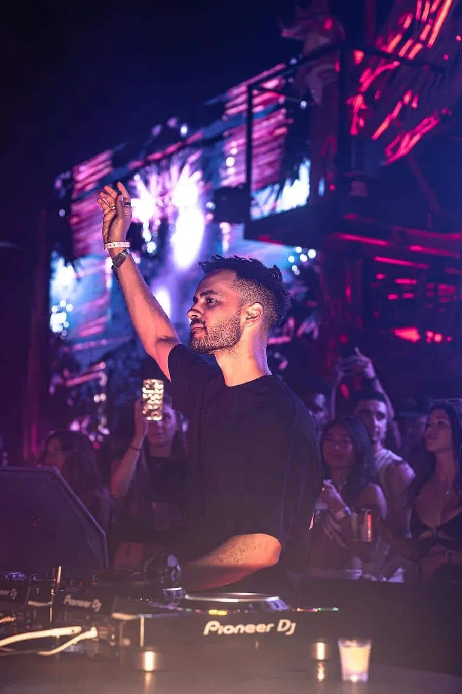 DJ performing at a dark nightclub, raising his right hand while mixing on a Pioneer DJ setup as a crowd watches.