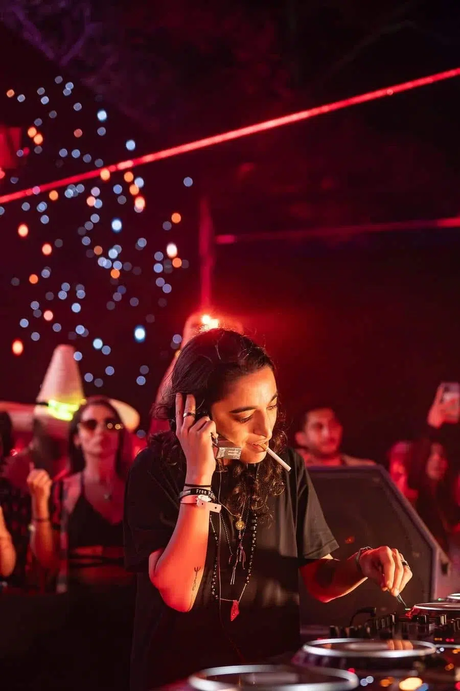 DJ woman in black adjusts a mixer at a nightclub, cigarette in mouth, with red stage lights and a cheering crowd in the background.