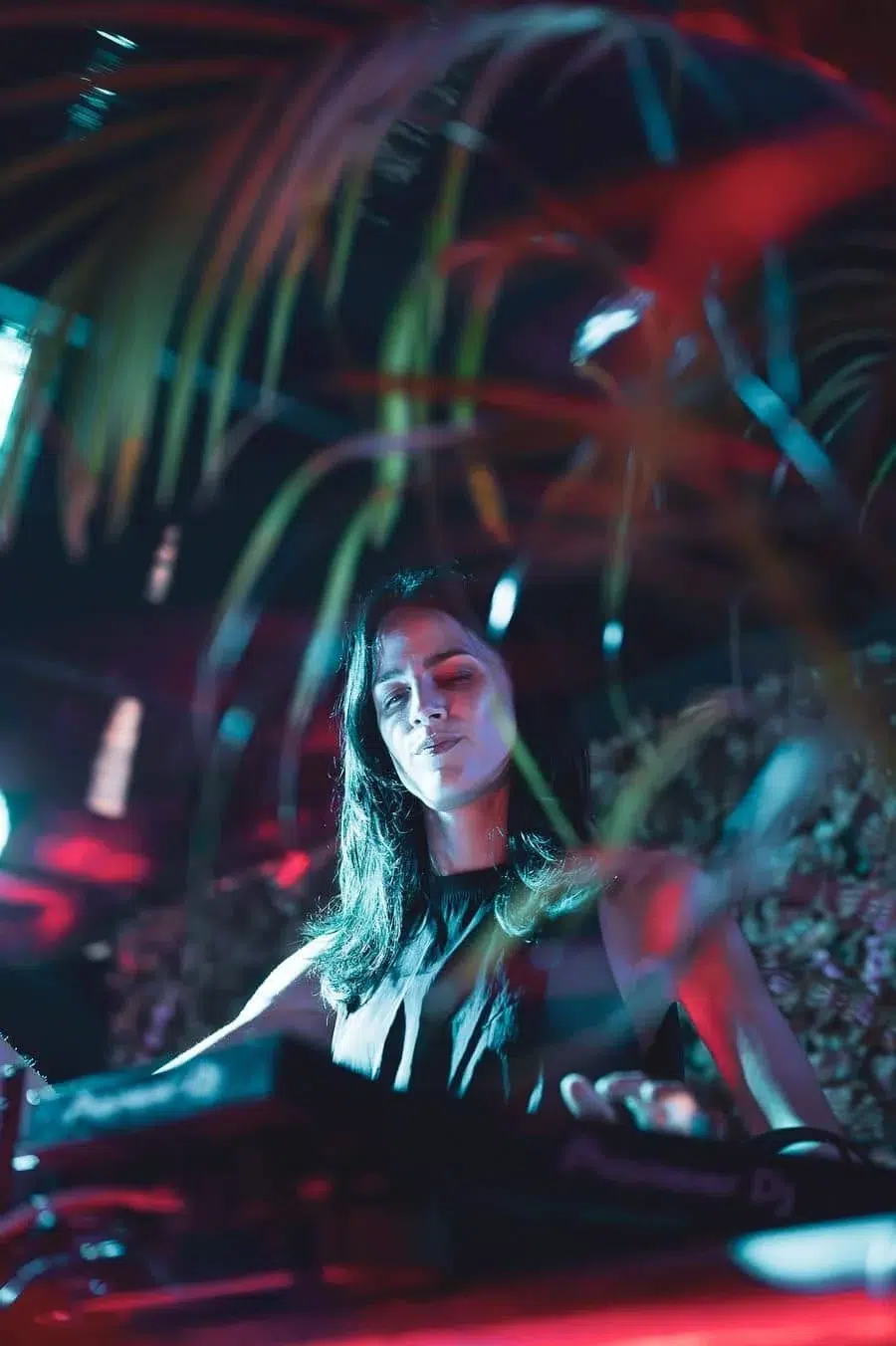 Female DJ smiling with eyes closed, mixing tracks under colorful nightclub lights against a tropical backdrop.
