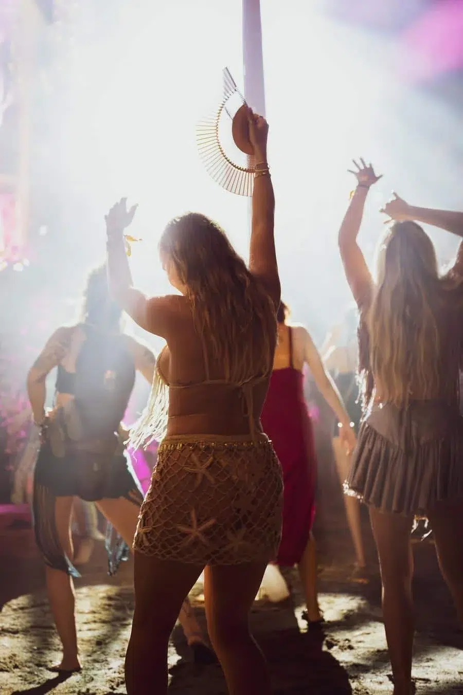Woman dancing at a nightclub, fringe shorts, raising a circular hat toward the ceiling amid bright lights and other partygoers.