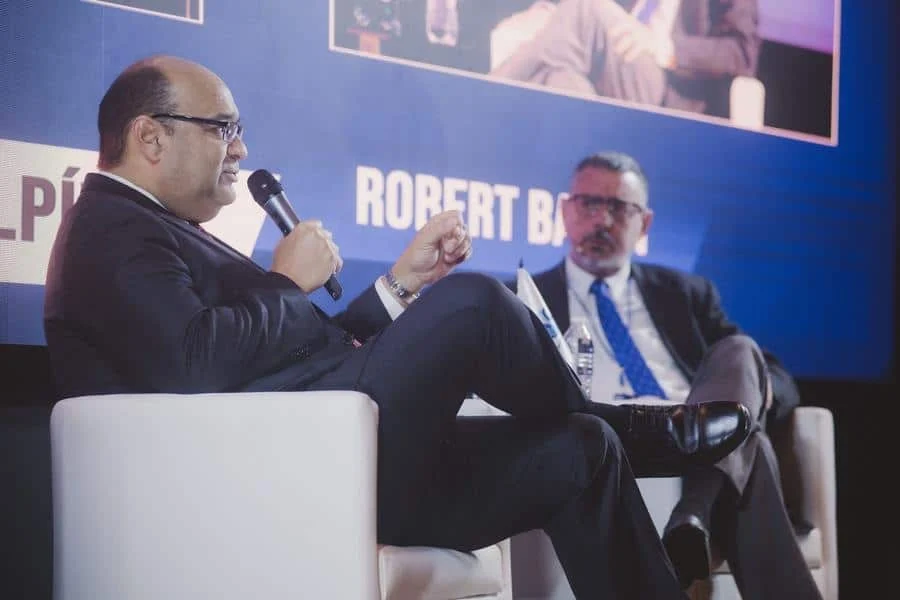 brands business and corporate photography -13 | Pacific Photography Man in a dark suit speaks into a handheld microphone on a panel stage, while another suited man listens in the background on a blue backdrop.