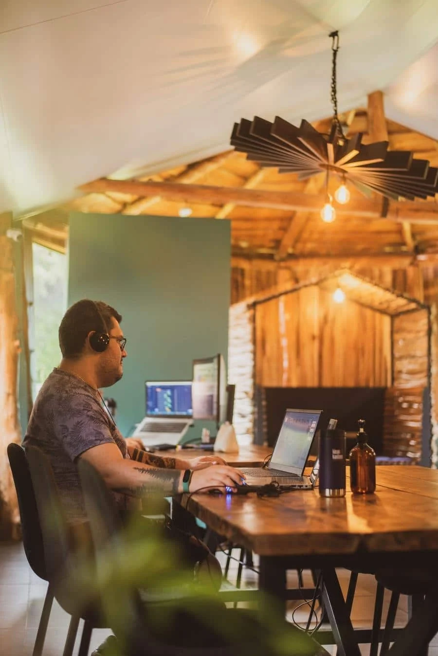 brands business and corporate photography -16 | Pacific Photography A man wearing headphones works on a laptop at a wooden communal desk in a rustic, warm-lit office or cafe setting.