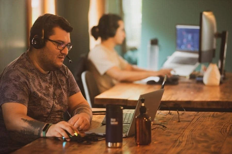 brands business and corporate photography -17 | Pacific Photography Support agent wearing a headset works at a wooden desk with a laptop, focused on the screen; another person at a computer is blurred in the background.