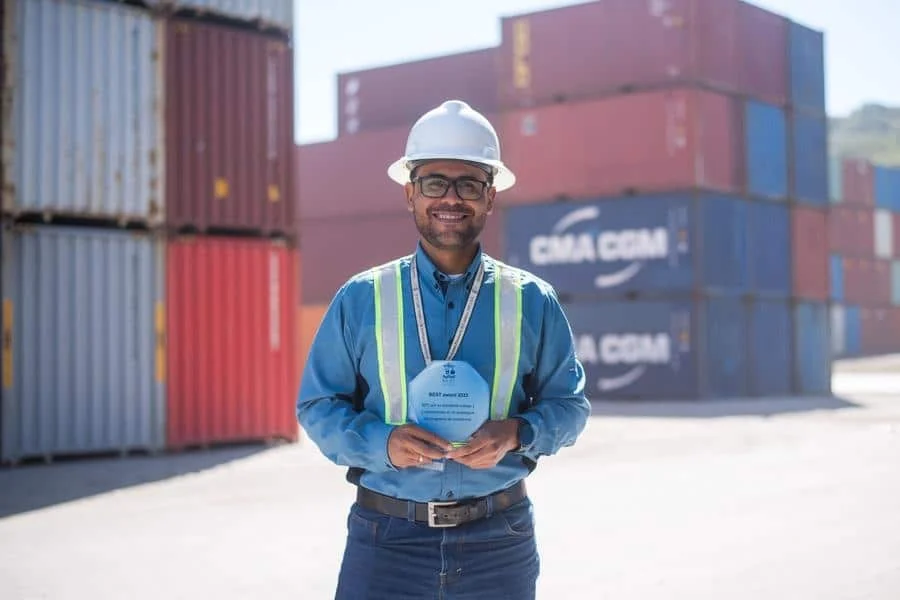 brands business and corporate photography -20 | Pacific Photography Construction worker in a hard hat and reflective vest holding a blue award at a container yard, smiling in the foreground.