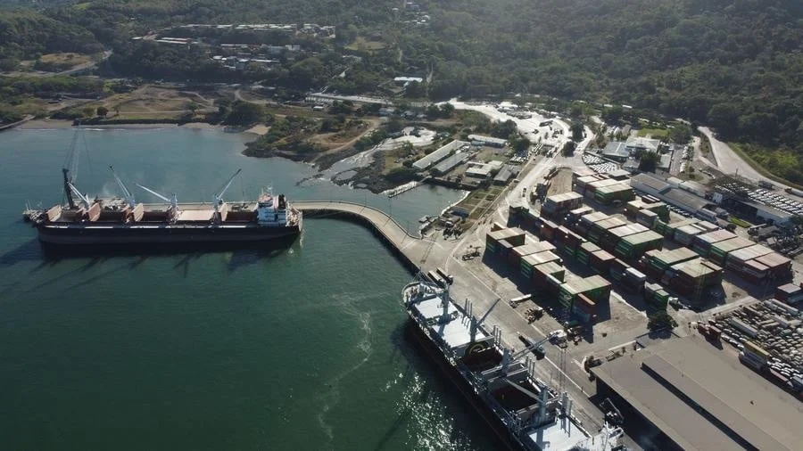 brands business and corporate photography -26 | Pacific Photography Aerial view of a shipping port with a large cargo ship at a curved pier and rows of shipping containers on the dock, surrounded by water and hills to the rear.
