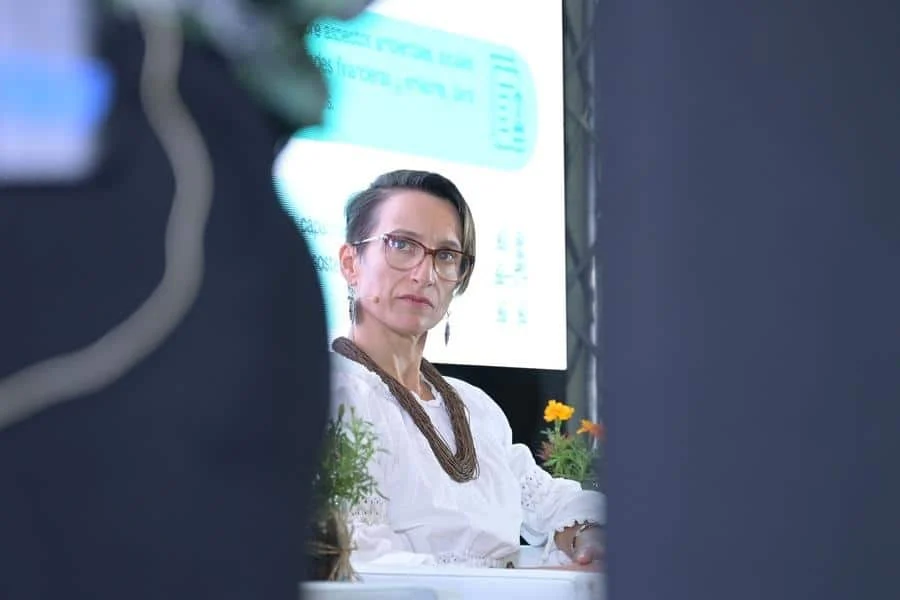 brands business and corporate photography -30 | Pacific Photography Woman with glasses and a brown beaded necklace sits at a conference panel, white blouse, plants nearby, projection screen in the background.