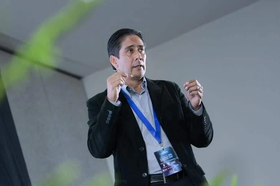 brands business and corporate photography -34 | Pacific Photography Male presenter in a blazer and blue lanyard badge speaking on stage, gesturing with his hands.