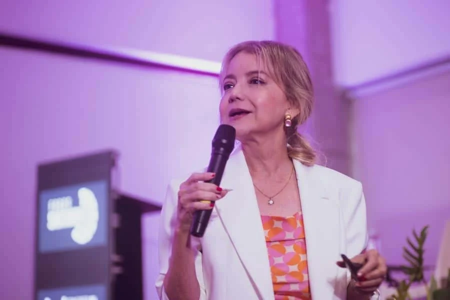 brands business and corporate photography -8 | Pacific Photography Female speaker on stage wearing a white blazer and orange patterned top, holding a microphone and speaking into it under purple lighting.