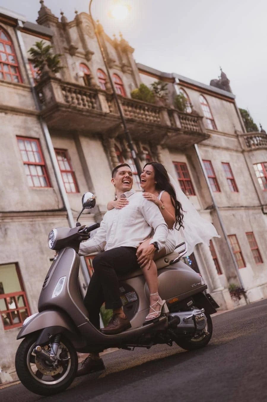 engagement photography costa rica-1 | Pacific Photography Couple on a gray scooter in front of a historic building; bride wearing white dress smiles as she hugs her partner.