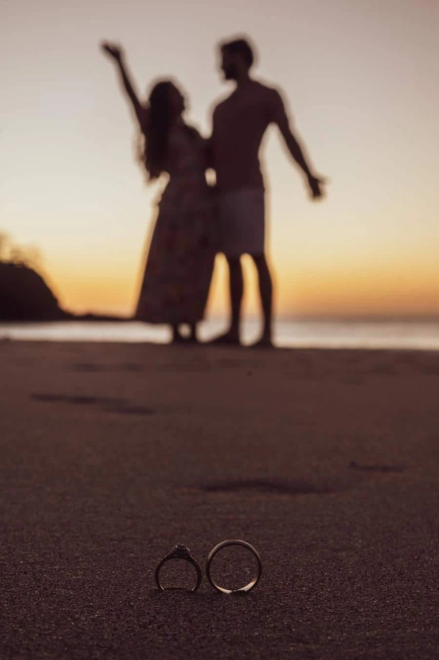 engagement photography costa rica-10 | Pacific Photography Silhouetted couple on a sunset beach, holding hands, with two rings on the sand in the foreground to symbolize engagement or marriage.