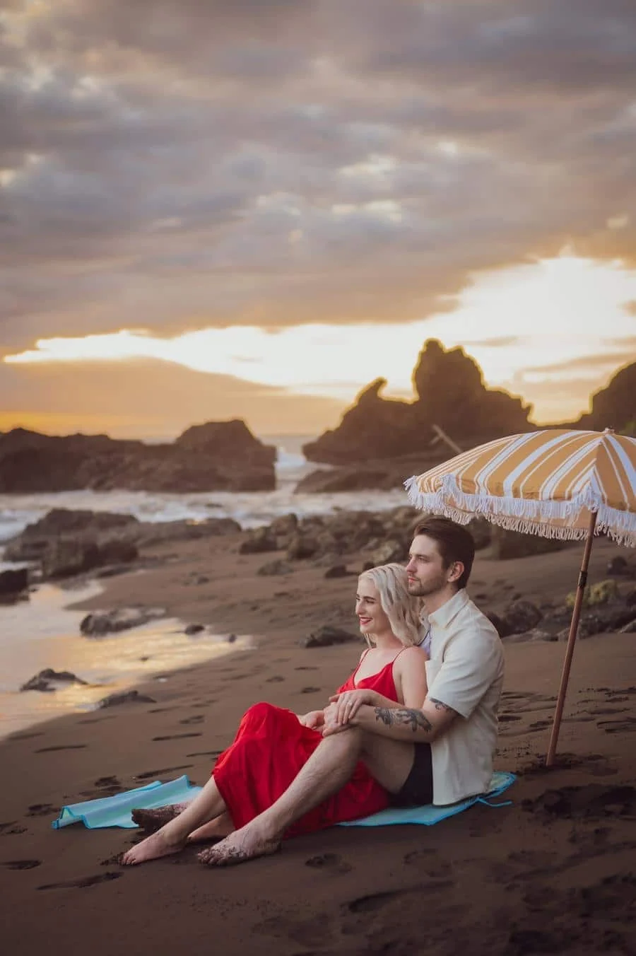 fotografía-de-compromiso-costa-rica-13 | Pacific Photography Pareja sentada en una playa de arena al atardecer bajo una sombrilla amarilla a rayas, tomados de la mano sobre una toalla azul.
