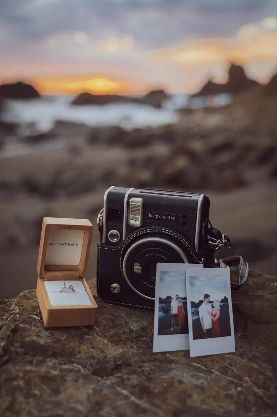 engagement photography costa rica-14 | Pacific Photography Black Instax mini 40 camera on a rocky shore beside an open wooden ring box with a diamond ring and two Polaroid photos at sunset by the sea